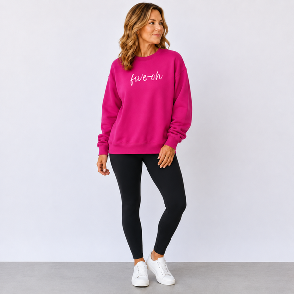Woman wearing a bright pink sweatshirt with 'five-oh' text, black leggings, and white sneakers on a light gray background.