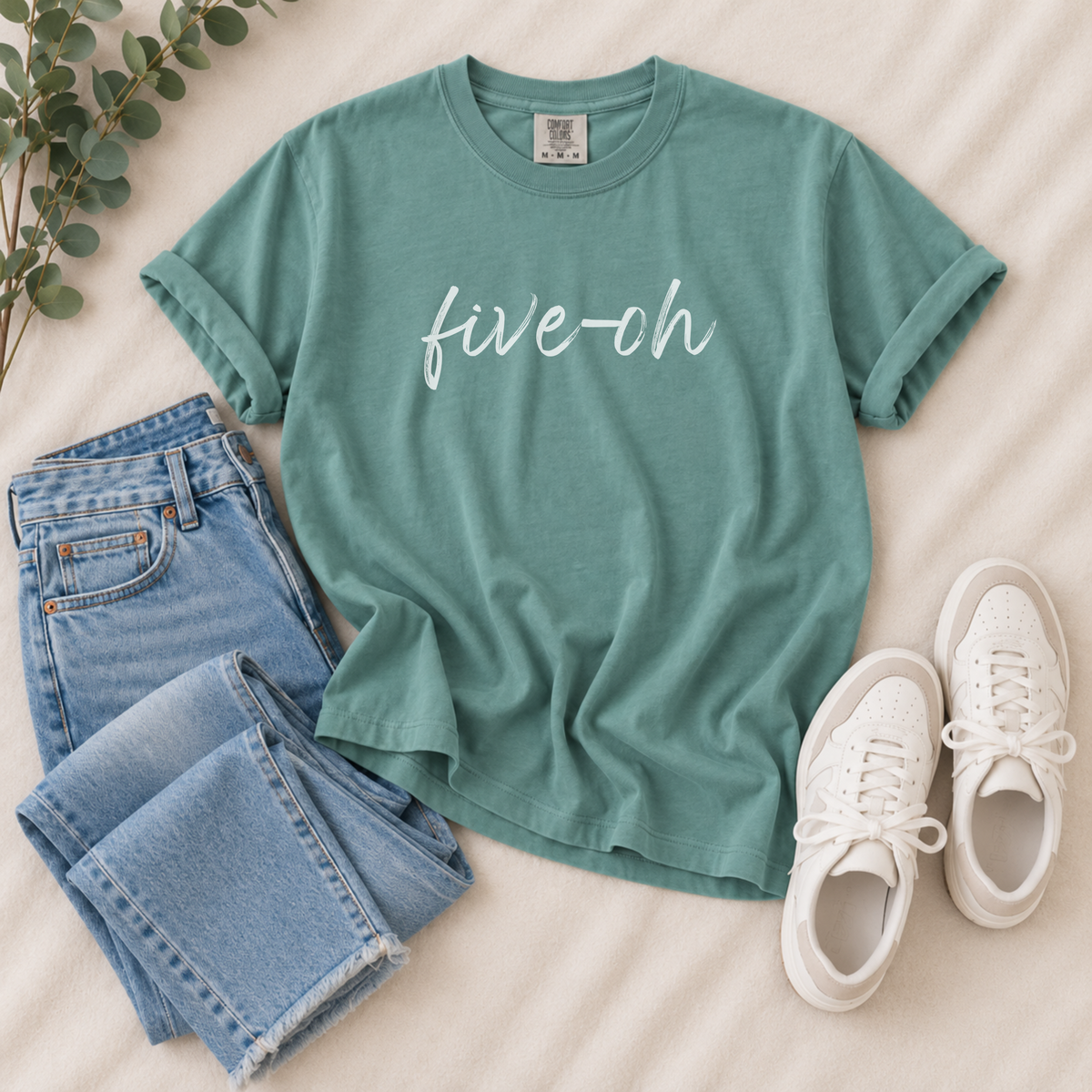 A styled flat-lay of a sage green t-shirt with 'five-oh' printed in white script across the chest, paired with light-wash blue jeans and white sneakers on a beige fabric background.
