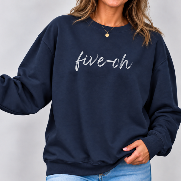 A woman wearing a navy blue crewneck sweatshirt with 'five-ch' written in white script across the chest, paired with light blue jeans against a neutral gray background.