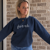 A woman wearing a navy blue crewneck sweatshirt with 'five-oh' embroidered in white script across the chest, standing against a brick wall.