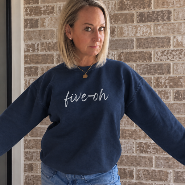 A woman wearing a navy blue crewneck sweatshirt with 'five-oh' embroidered in white script across the chest, standing against a brick wall.