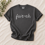 A dark charcoal gray t-shirt with the phrase 'five-oh' printed in white cursive across the chest, laid flat on a beige fabric background with a decorative eucalyptus branch in the top-left corner.