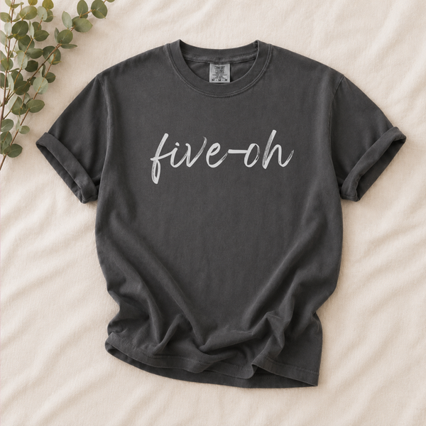 A dark charcoal gray t-shirt with the phrase 'five-oh' printed in white cursive across the chest, laid flat on a beige fabric background with a decorative eucalyptus branch in the top-left corner.