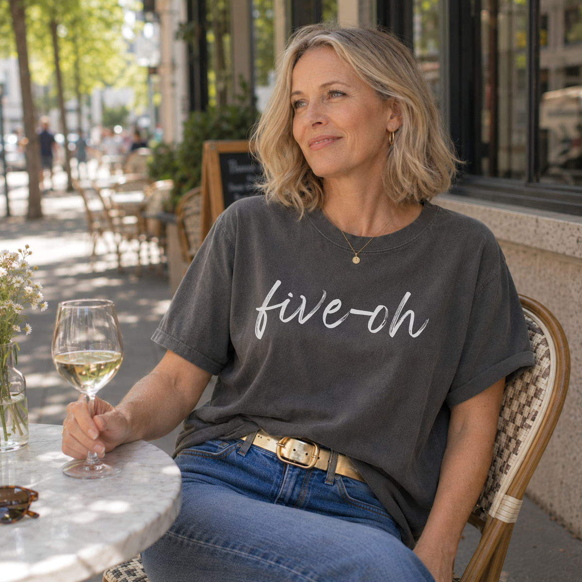 Woman sitting at an outdoor cafe holding a glass of wine, wearing a 'five-oh' t-shirt.