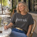 Woman sitting at an outdoor cafe holding a glass of wine, wearing a 'five-oh' t-shirt.