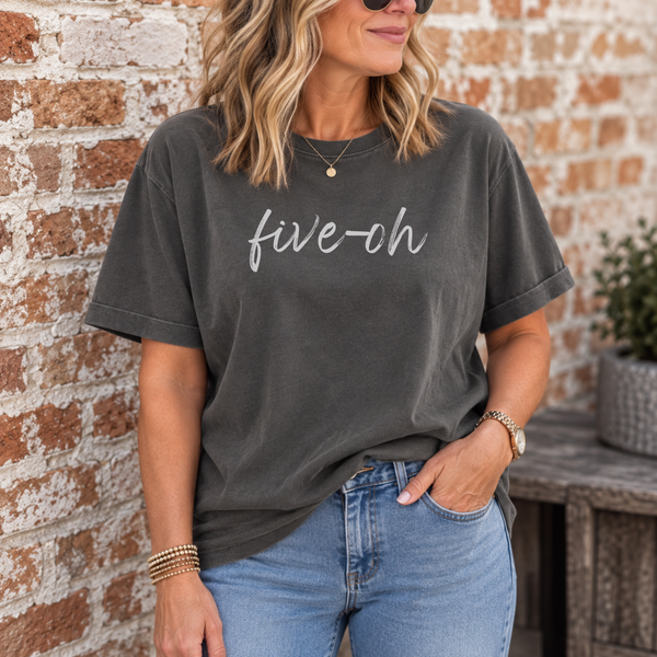 A woman wearing a dark charcoal gray t-shirt with the text 'five-oh' printed in white script across the chest, paired with light blue jeans, standing against a rustic brick wall.