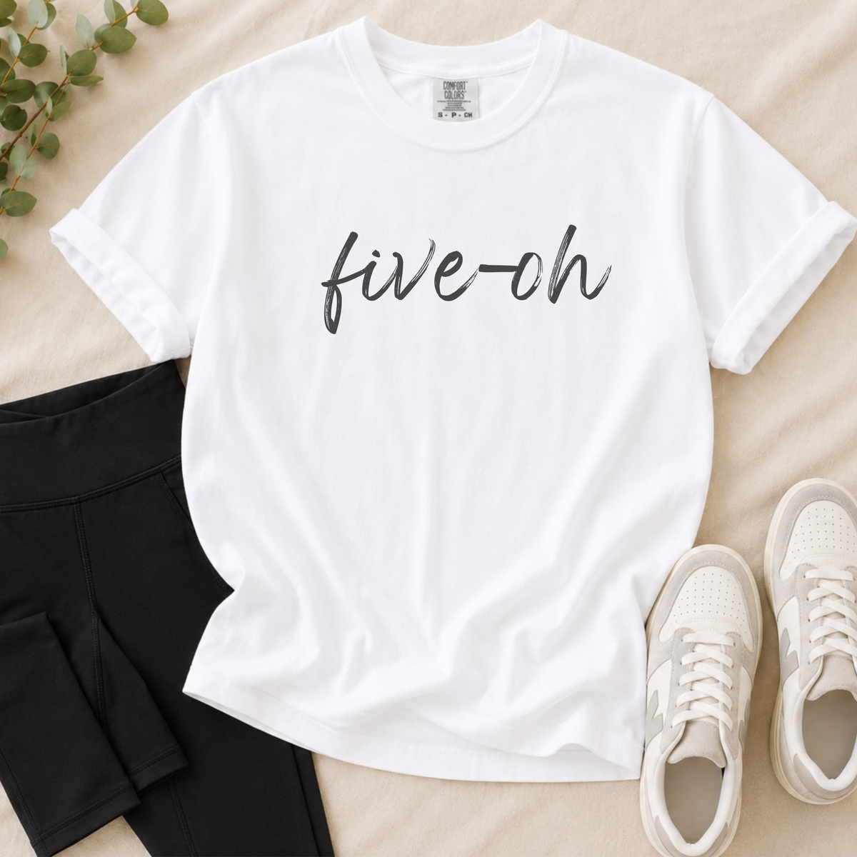A white short-sleeve T-shirt with the text 'five-oh' printed in black script across the chest, styled in a flat lay with black shorts, white sneakers, and a small green plant on a beige fabric background.