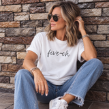 A woman wearing a white graphic t-shirt with 'five-ch' printed in black script, paired with light blue jeans and white sneakers, sitting against a rustic stone wall.
