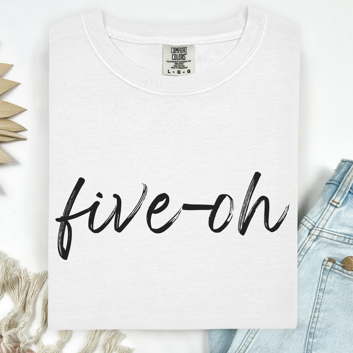 A folded Comfort Colors white t-shirt with 'five-oh' text on a white background, sitting next to a pair of jeans.