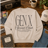 A cream-colored crewneck sweatshirt with a black graphic print reading 'GEN X Social Club EST. 1965-1980 *we don't actually meet' is displayed in a flat-lay arrangement on a wooden table surrounded by vintage props including a cassette tape, a coffee mug, a notebook, and jeans.