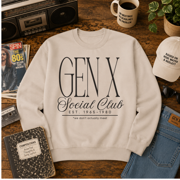 A cream-colored crewneck sweatshirt with a black graphic print reading 'GEN X Social Club EST. 1965-1980 *we don't actually meet' is displayed in a flat-lay arrangement on a wooden table surrounded by vintage props including a cassette tape, a coffee mug, a notebook, and jeans.