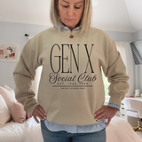 A woman wearing a beige crewneck sweatshirt with 'GEN X Social Club EST. 1965-1980' printed in black on the front, layered over a blue and white striped button-down shirt in a bedroom setting.