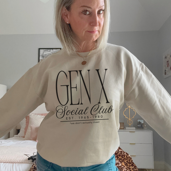 A woman wearing a beige crewneck sweatshirt with 'GEN X Social Club EST. 1968-1980' printed in black on the front, standing in a bedroom setting.