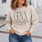 A woman wearing a beige crewneck sweatshirt with a black graphic print that reads 'GEN X Social Club EST. 1965-1980' and 'we don't actually meet', paired with blue jeans in an outdoor setting.