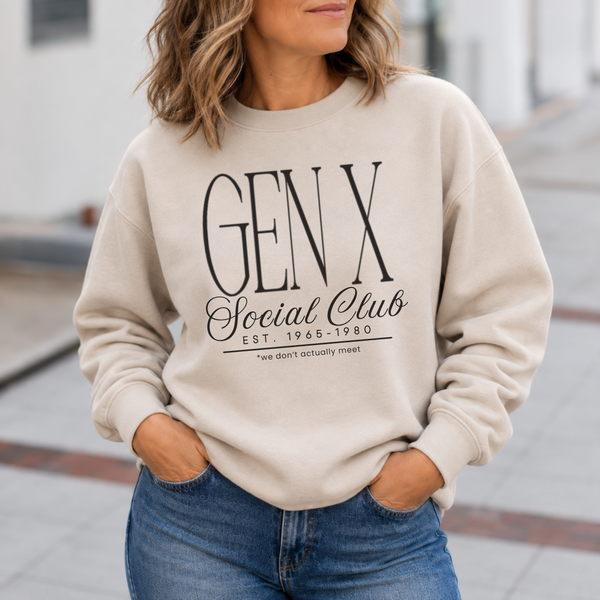 A woman wearing a beige crewneck sweatshirt with a black graphic print that reads 'GEN X Social Club EST. 1965-1980' and 'we don't actually meet', paired with blue jeans in an outdoor setting.