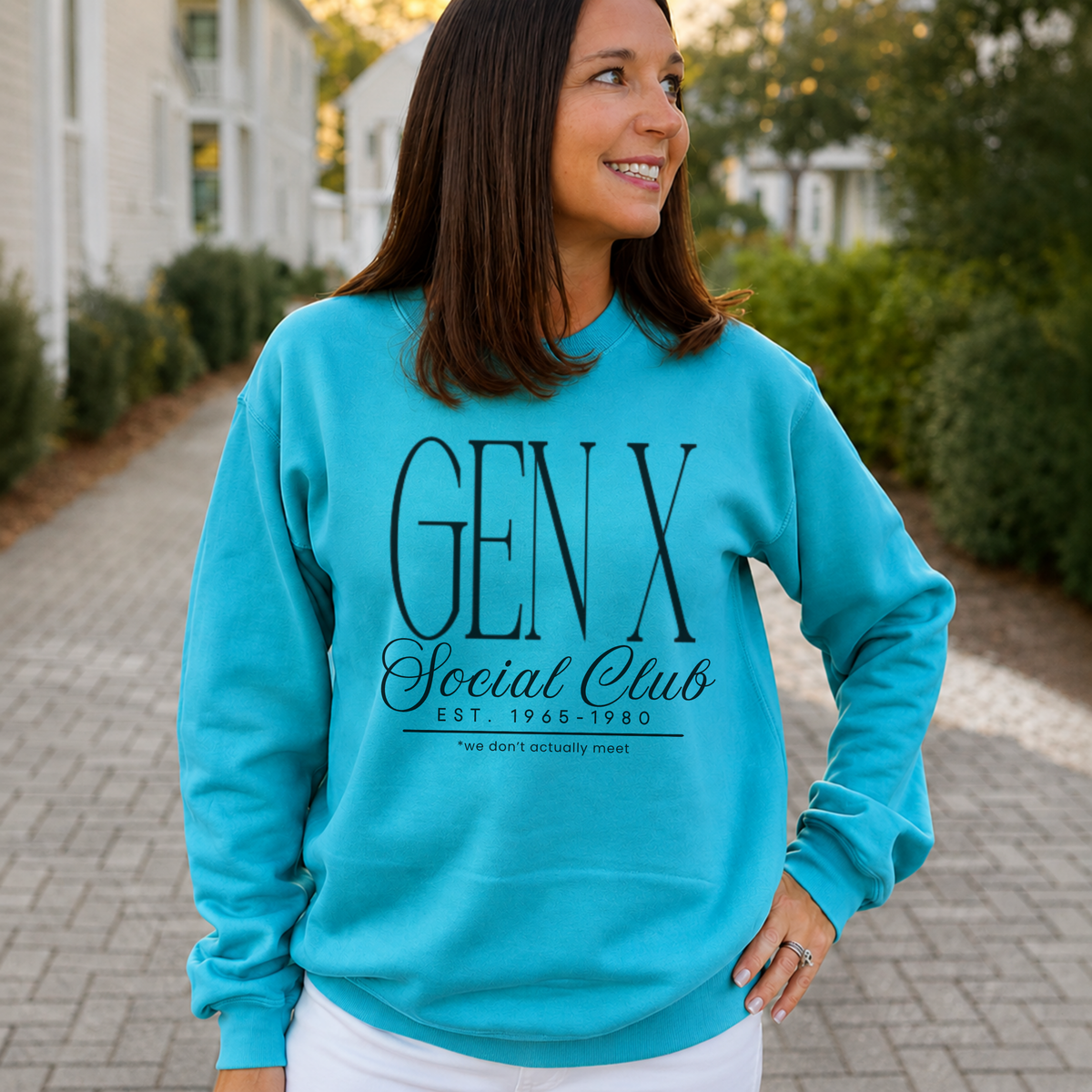 A woman wearing a bright turquoise crewneck sweatshirt with a black graphic print that reads 'GEN X Social Club EST. 1968-1980' and 'the next generation twist', standing outdoors on a paved walkway with blurred houses and greenery in the background.