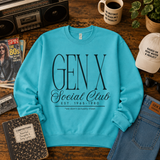 A turquoise crewneck sweatshirt with 'GEN X Social Club' printed in black on the front, styled in a retro-themed flat lay on a wooden table with a cassette tape, coffee mug, notebook, and denim accessories.
