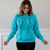 A woman wearing a bright turquoise crewneck sweatshirt with 'GEN X Social Club' printed in black on the front, paired with black pants, standing against a light gray wall with white trim.