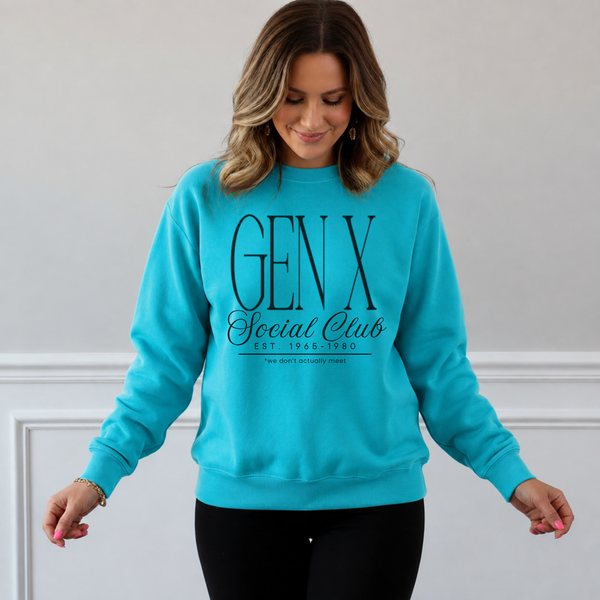 A woman wearing a bright turquoise crewneck sweatshirt with 'GEN X Social Club' printed in black on the front, paired with black pants, standing against a light gray wall with white trim.
