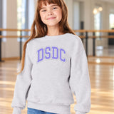 A young girl with long brown hair smiling in a light gray sweatshirt with 'DSDC' printed in purple and white arched letters on the chest, set against a blurred indoor studio background.
