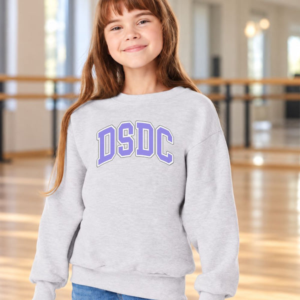 A young girl with long brown hair smiling in a light gray sweatshirt with 'DSDC' printed in purple and white arched letters on the chest, set against a blurred indoor studio background.