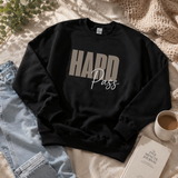 A black crewneck sweatshirt laid flat on a cream fabric surface, styled with light blue jeans, a white mug of coffee, a book, and a small plant. The sweatshirt has large taupe-gray block letters reading “HARD” and smaller white script text reading “Pass” across the chest.
