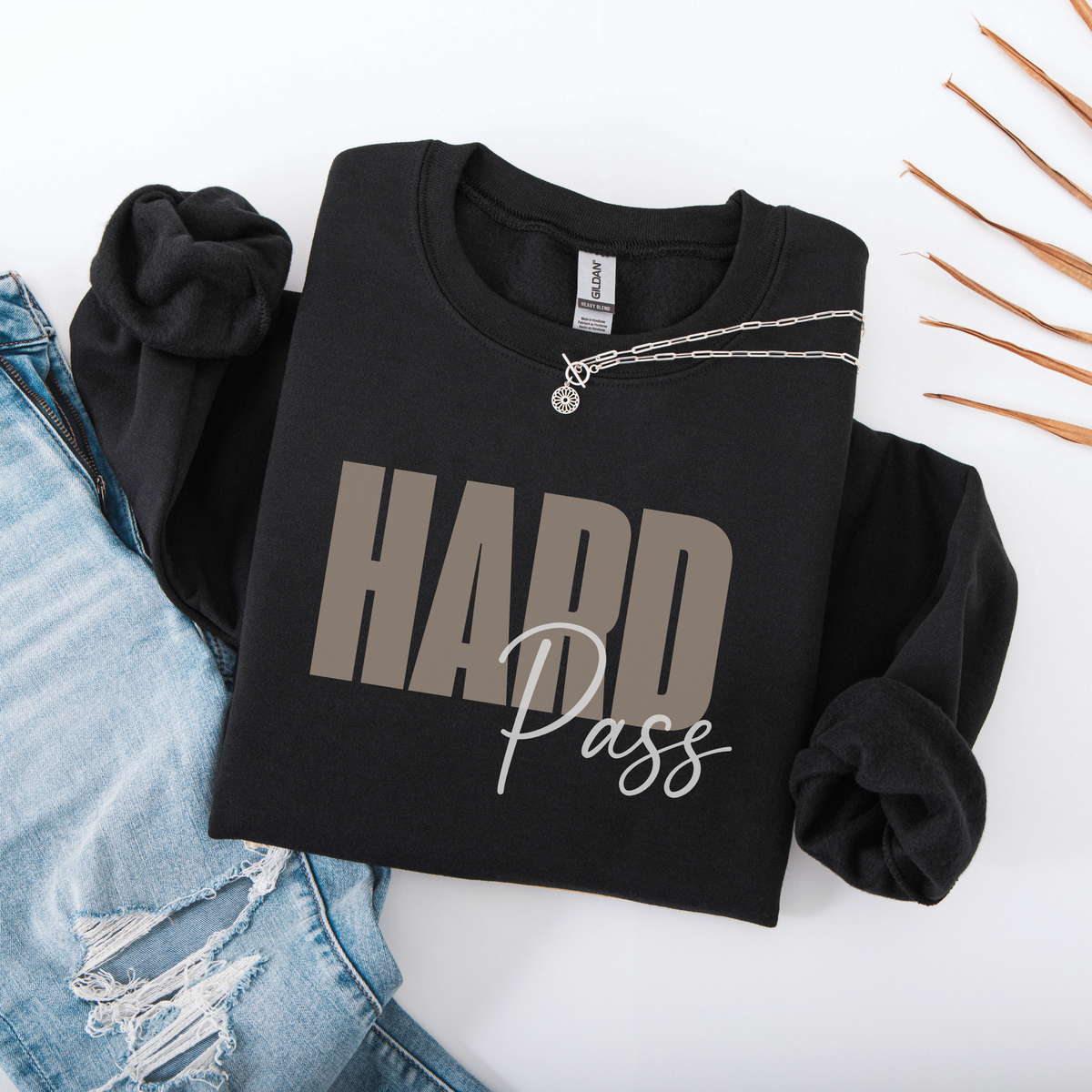 A black long-sleeve sweatshirt with 'HARD Pass' printed on the front in beige and white, styled in a flat lay with light blue jeans, a silver necklace, and dried orange leaves on a white background.