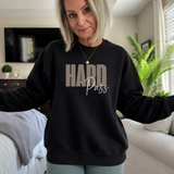 A woman wearing a black crewneck sweatshirt with 'HARD Pass' printed on the front, standing in a living room setting.