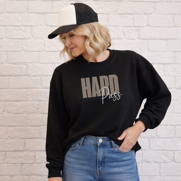 A woman wearing a black crewneck sweatshirt with 'HARD Pass' printed on the front in gray and white, paired with blue jeans and a black-and-white trucker hat, standing against a white brick wall.