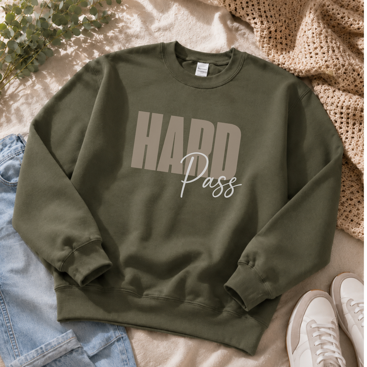 An olive green crewneck sweatshirt with 'HARD Pass' printed on the front, styled in a flat lay with light blue jeans, white sneakers, a beige knit blanket, and green leaves on a beige fabric background.