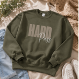 An olive green crewneck sweatshirt with 'HARD Pass' printed on the front, styled in a flat lay with light blue jeans, white sneakers, a beige knit blanket, and green leaves on a beige fabric background.