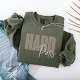 An olive green crewneck sweatshirt with rolled sleeves, featuring a graphic print that reads 'HARD Pass' in beige and white. The sweatshirt is styled in a flat-lay arrangement with light-wash distressed denim jeans and a silver chain necklace on a white background.