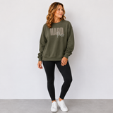 A woman modeling an olive green crewneck sweatshirt with 'HARD Pass' printed in gray across the chest, paired with black leggings and white sneakers, standing against a light gray wall.