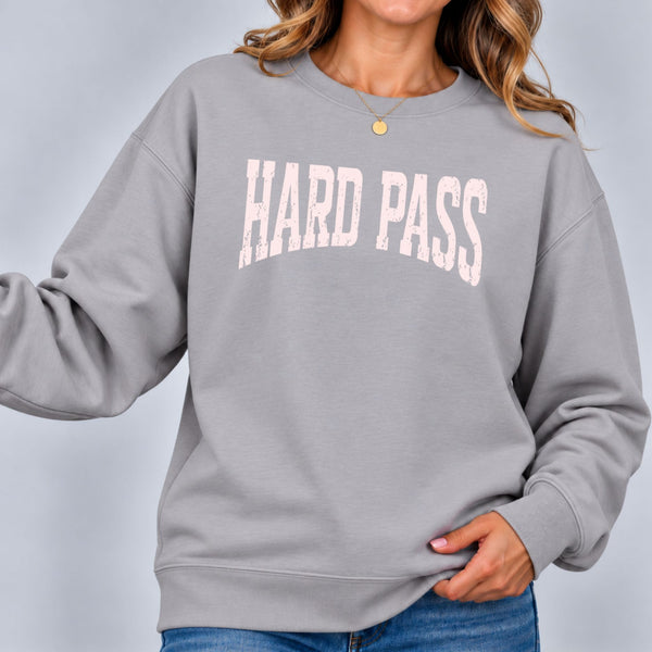 Woman wearing the stone gray Hard Pass sweatshirt with light pink lettering, paired with blue jeans, in a close-up studio shot.