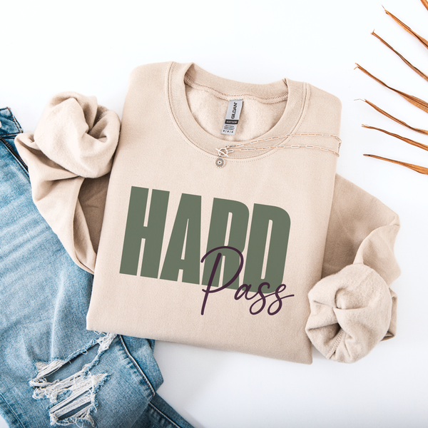 A folded beige sweatshirt with rolled sleeves, featuring the text 'HARD Pass' printed on the front. The sweatshirt is styled in a flat lay with a pair of distressed blue jeans on a white background.