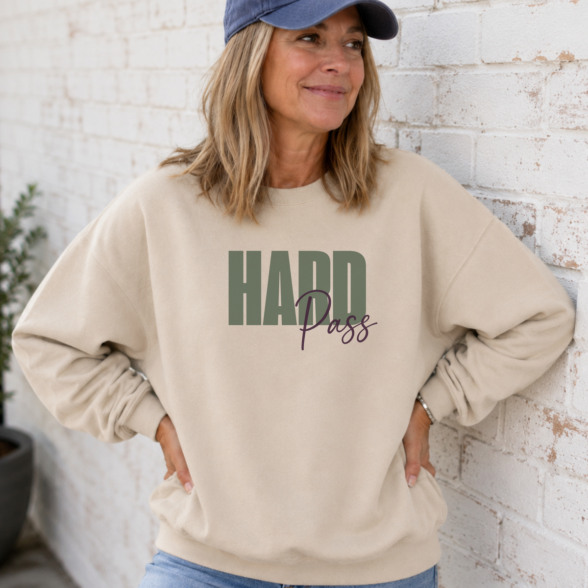 A woman wearing a beige crewneck sweatshirt with 'HARD Pass' printed on the front, paired with a navy blue baseball cap and blue jeans, leaning against a white brick wall.