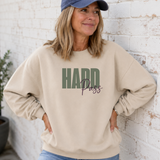 A woman wearing a beige crewneck sweatshirt with 'HARD Pass' printed on the front, paired with a navy blue baseball cap and blue jeans, leaning against a white brick wall.