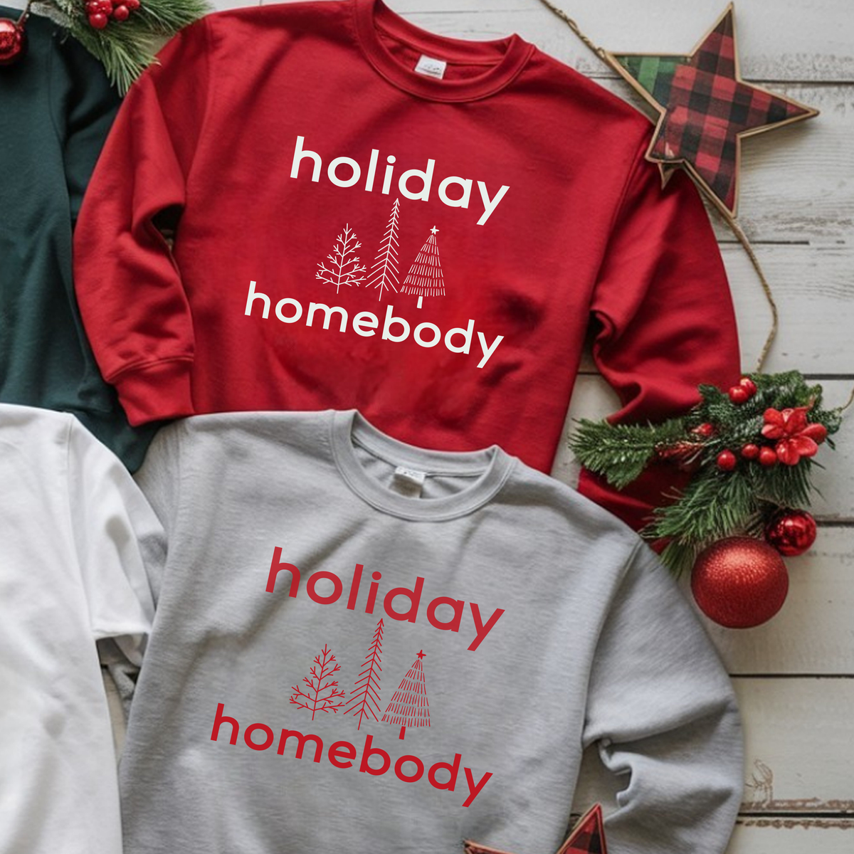 Red and gray sweatshirts with 'holiday homebody' text and Christmas decorations on a wooden surface.