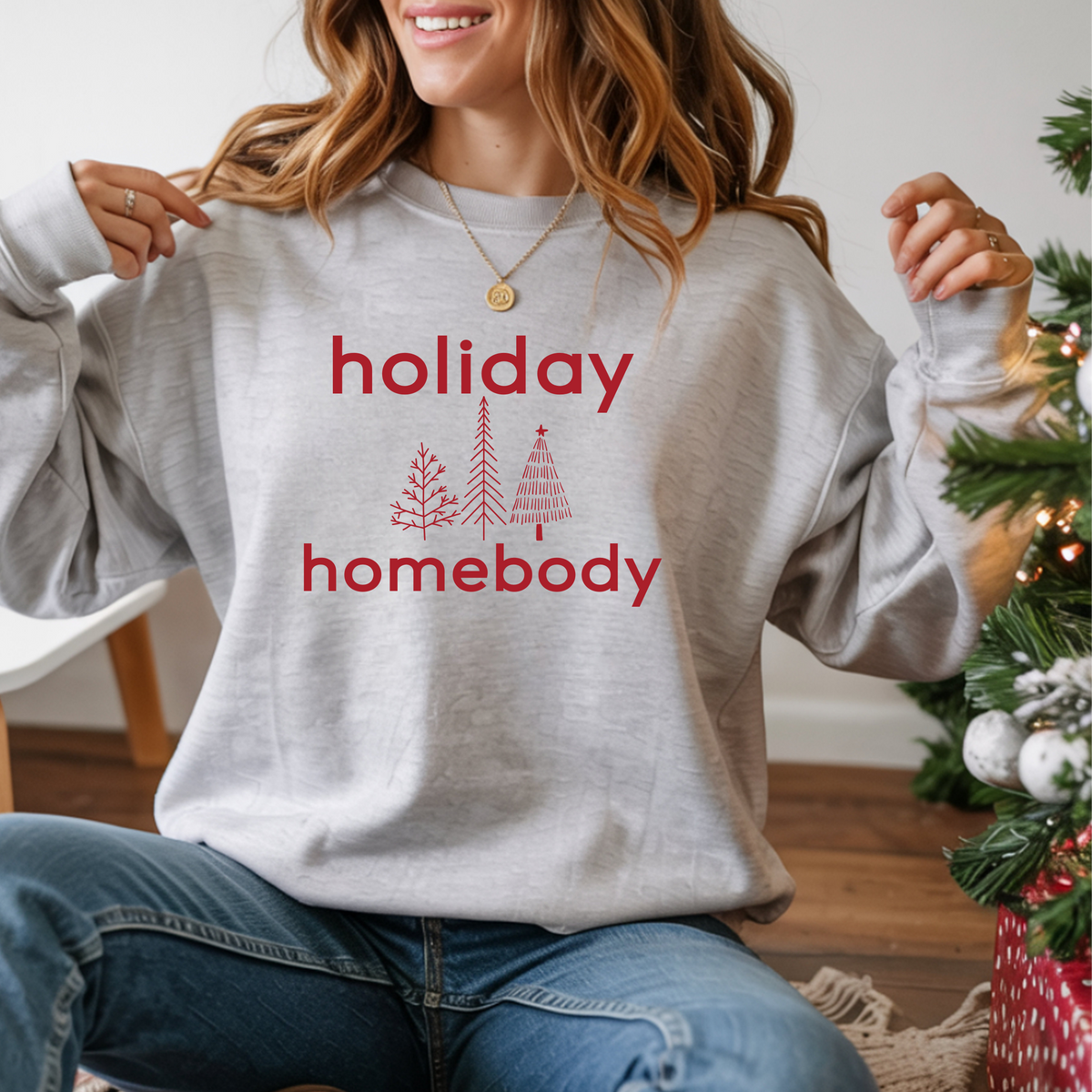 Person wearing a gray sweatshirt with 'holiday homebody' text and Christmas tree graphics, sitting in front of a decorated Christmas tree.