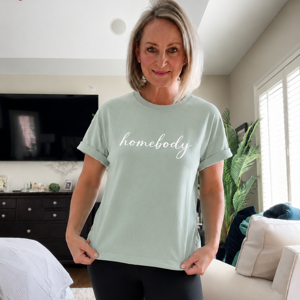 A woman standing in a bedroom offee with a light green shirt on that says "homebody" in script writing.