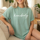 Woman wearing a bay green t-shirt with 'homebody' text, sitting on a couch in a cozy living room.
