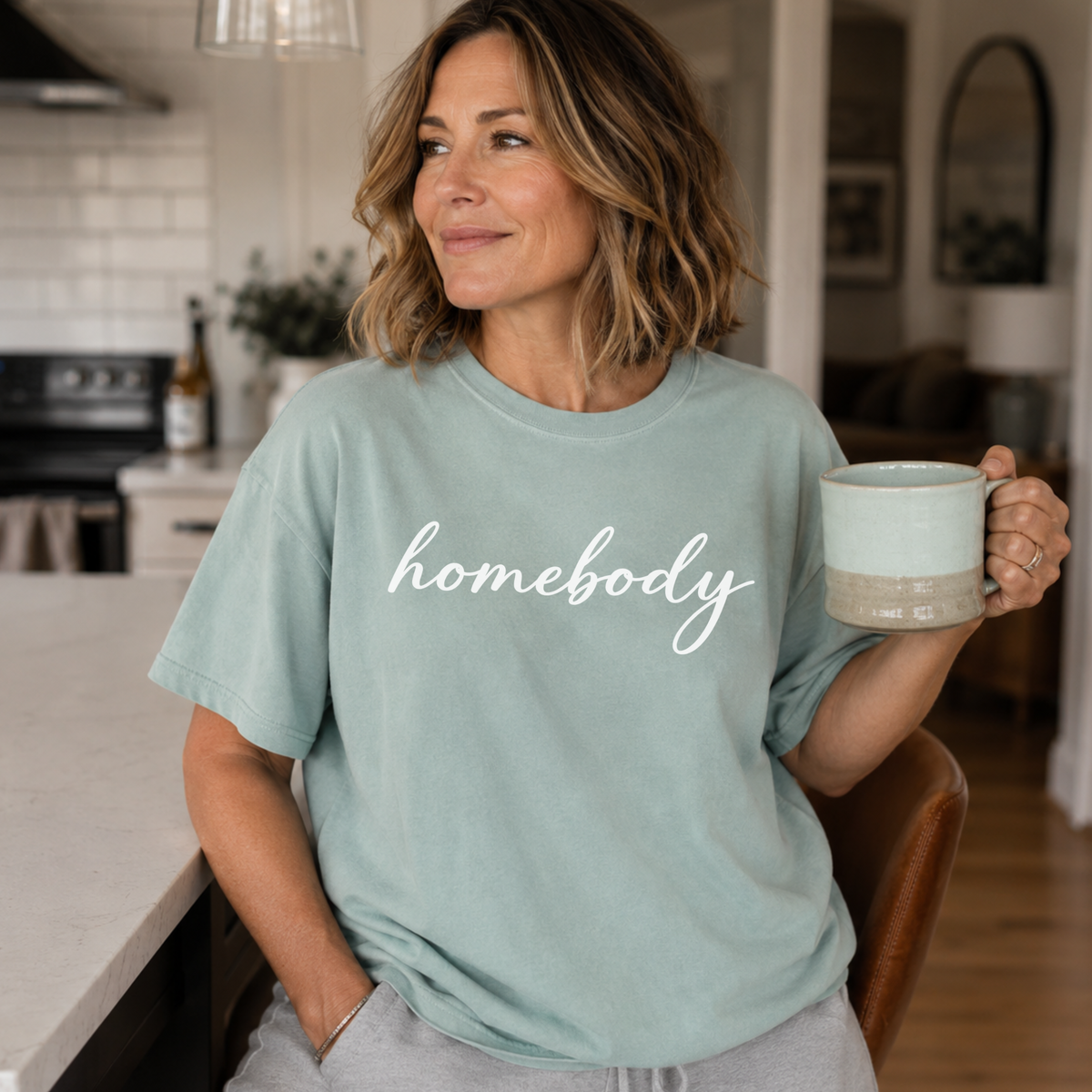 A woman standing in a kitchen holding coffee with a light green shirt on that says "homebody" in script writing.