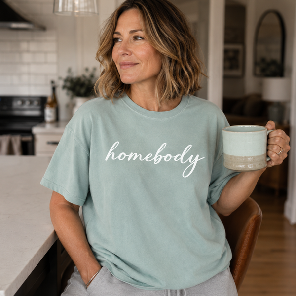A woman standing in a kitchen holding coffee with a light green shirt on that says "homebody" in script writing.