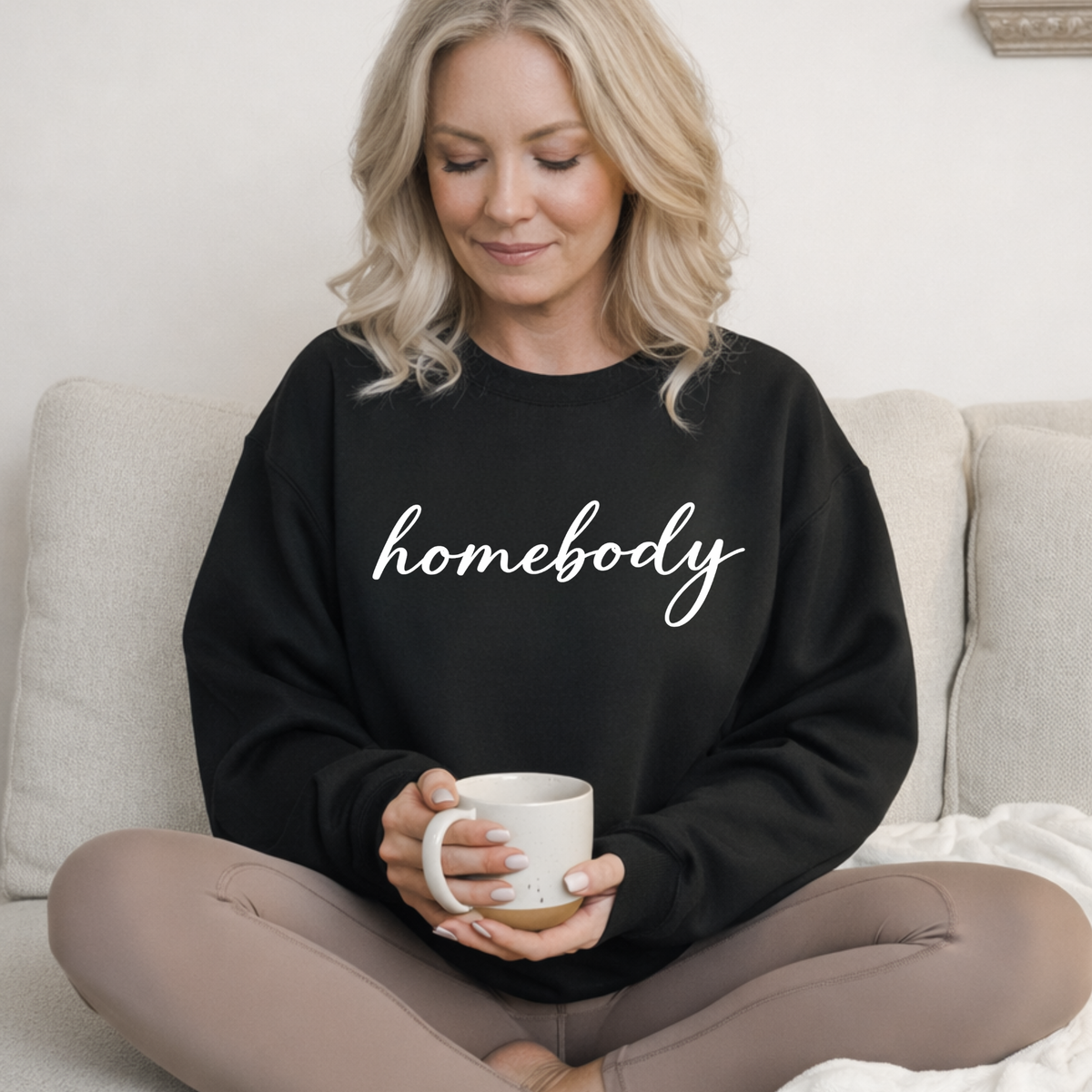 Woman wearing a black sweatshirt with 'homebody' text, holding a mug on a couch.