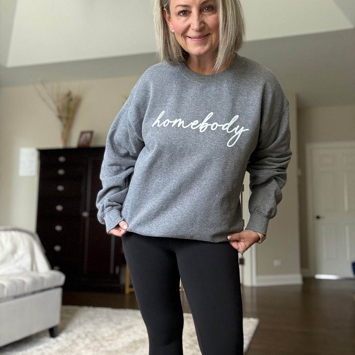 Woman wearing a gray sweatshirt with 'homebody' text in a living room.