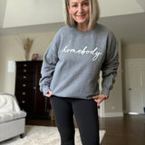 Woman wearing a gray sweatshirt with 'homebody' text in a living room.