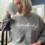 A woman wearing a gray sweatshirt with 'homebody' text, holding a mug indoors.