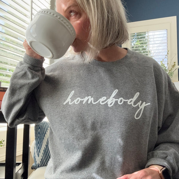 A woman wearing a gray sweatshirt with 'homebody' text, holding a mug indoors.