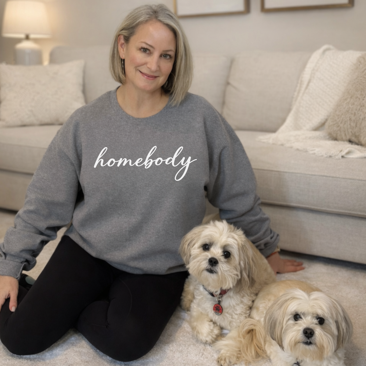Woman wearing a 'homebody' sweatshirt with two dogs on a carpeted floor.