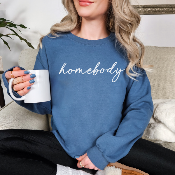 Person wearing a blue sweatshirt with 'homebody' text, holding a white mug.